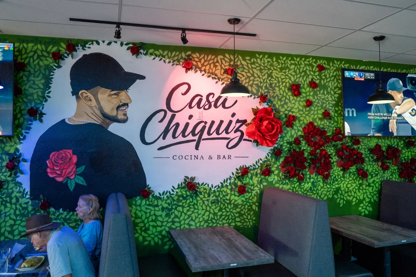 Casa Chiquiz logo mural with red roses on green hedge wall over booths